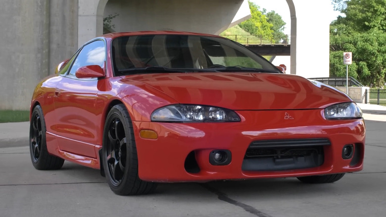 The front three-quarter angle of a red second-generation Mitsubishi Eclipse GSX coupe parked on pavement, featuring the late-model aggressive front fascia, side skirts, and black multi-spoke wheels.