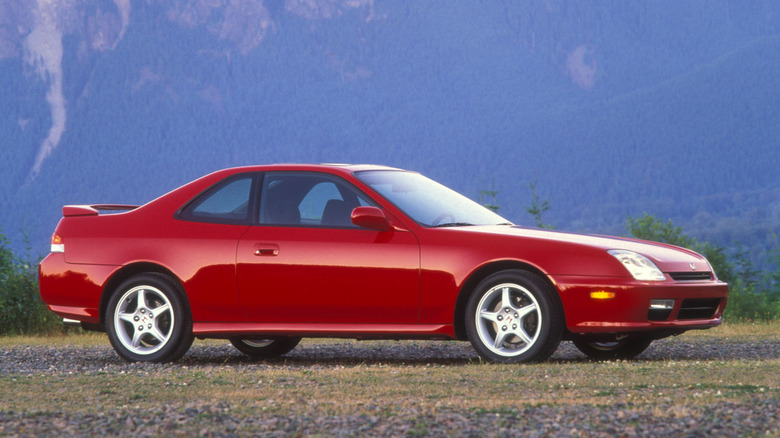 A side profile view of a red fifth-generation Honda Prelude Type SH parked on gravel against a mountainous backdrop, highlighting its crisp coupe silhouette, factory rear spoiler, and five-spoke alloy wheels.