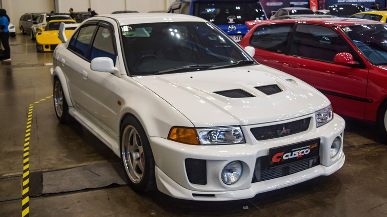 The front three-quarter of a white Mitsubishi Lancer Evolution V in a car garage filled with Japanese cars