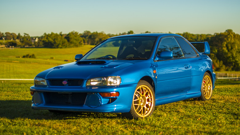 The front three-quarter view of a Sonic Blue Subaru Impreza 22B STi coupe parked on a sunlit grassy field, highlighting its blistered widebody fenders, hood scoop, and gold BBS multi-spoke wheels.