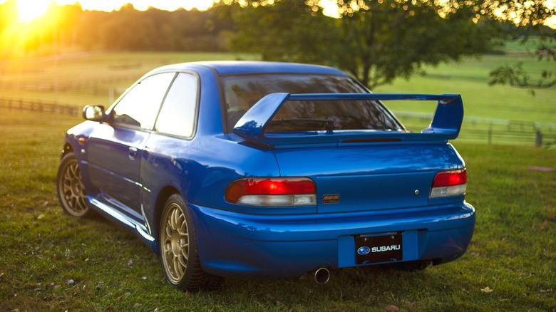 The rear three-quarter view of a blue Subaru Impreza 22B STi coupe on grass at sunset, showing its tall factory rear wing, wide rear quarter panels, and single exhaust tip
