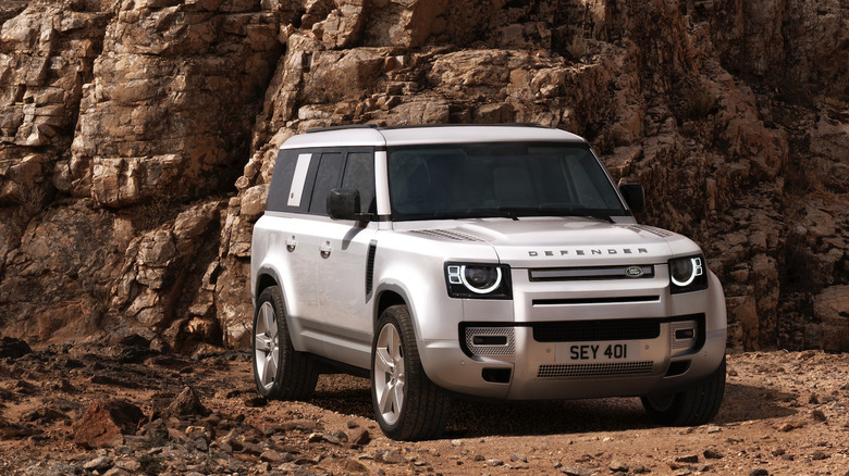 A Land Rover Defender 130 sits on dirt near cliffs.