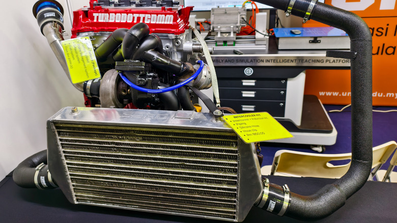 This is an intercooler for a turbocharger on a desk beside a 4cyl turbocharged engine with a red valve cover.