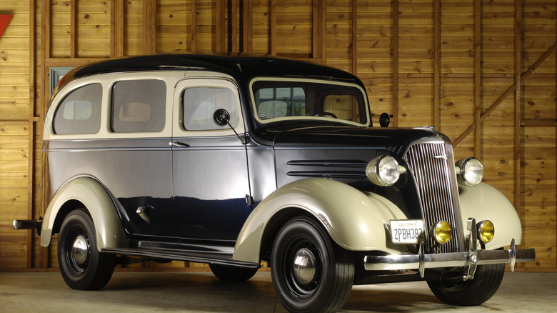 A achromatic and tan 1930s-era two-door Chevrolet Suburban parked successful a woody building.
