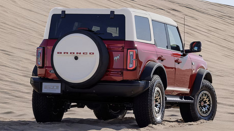 A red Bronco with a rear spare sits in sand in front of a large dune.