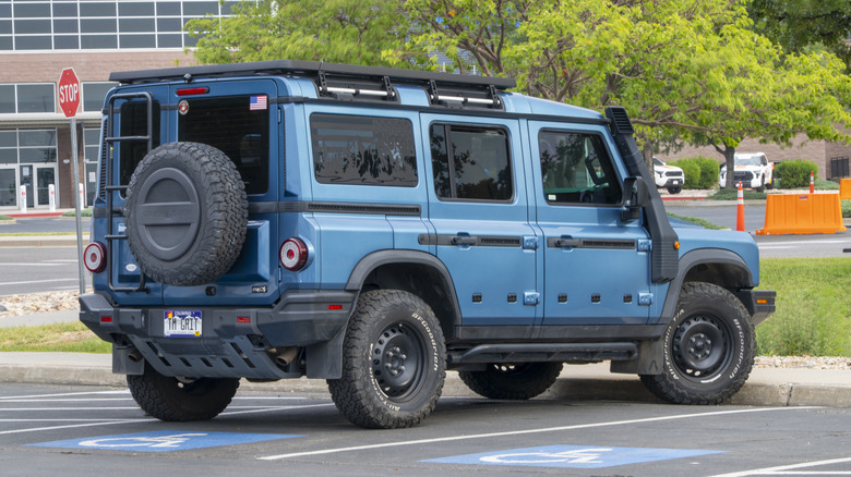 A blue Ineos Grenadier sits parked in a lot with trees, a stop sign, and an office building in the background