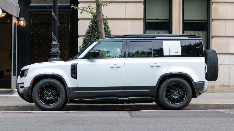 A white Land Rover Defender sits parked on the street in front of a stone building