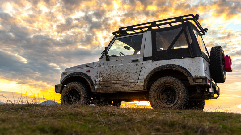 A white, mud-splattered Suzuki Samurai sits parked under lovely skies.