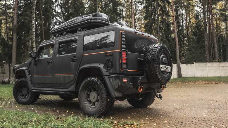 A black Hummer sits on a cobblestone driveway with a wall and trees in the background