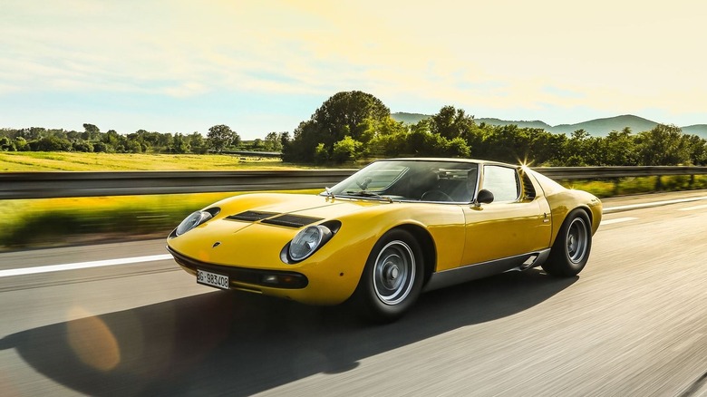 A dynamic rolling front 3/4 shot of a classic bright yellow Lamborghini Miura driving on a multi-lane highway during a bright, sunny day.