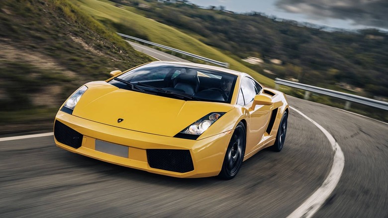A dynamic, front 3/4 action shot of a bright yellow Lamborghini Gallardo speeding along a curved, two-lane asphalt road.