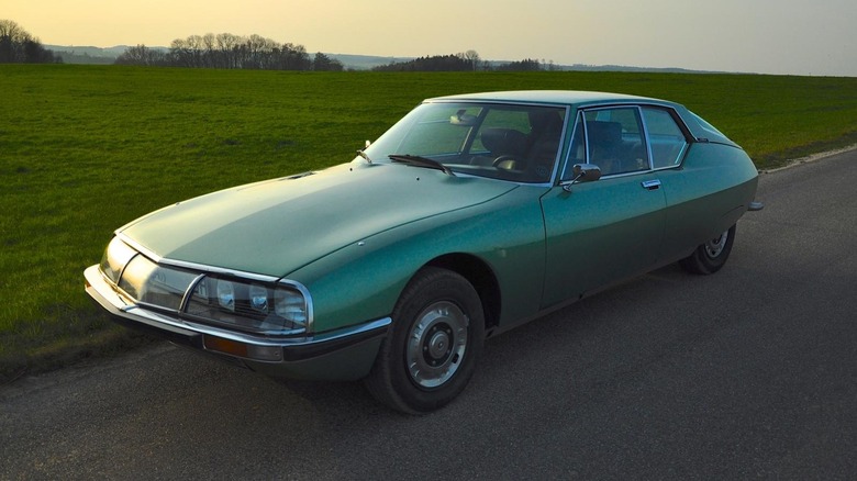 A front-three-quarter view of a classic metallic jade-green Citroën SM parked on the shoulder of a quiet asphalt road.
