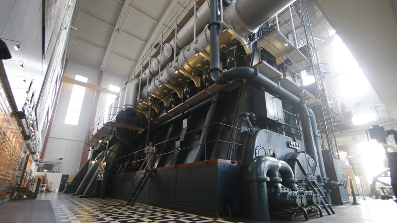 A perspective shot from the floor of the DieselHouse museum in Copenhagen, looking up at the world-famous B&W 884WS-150. The massive, four-story-tall black engine block is surrounded by intricate silver piping and industrial gantries.