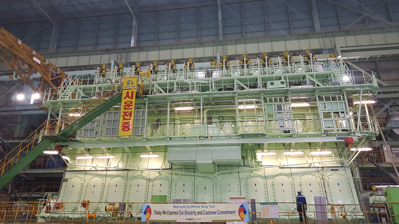 An immense, pale-green 11-cylinder MAN B&W G95ME engine inside a massive shipbuilding workshop. The engine is several stories high, featuring a "Welcome to Official Shop Test" banner at its base. A worker stands right to the banner, showcasing the engine's immense size.