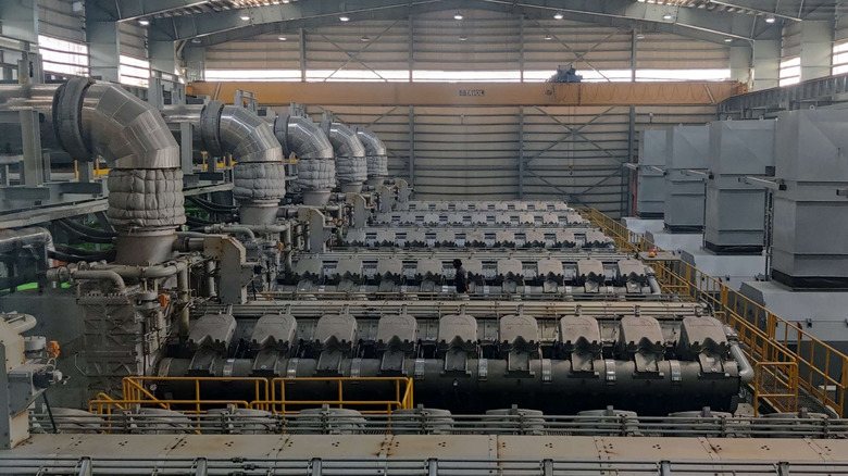 A high-angle interior shot of a massive industrial engine hall or power plant featuring a row of MAN 18V48/60TS V18 dieselengines.