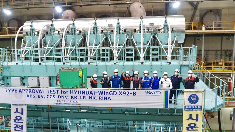 A wide shot of a massive teal-colored WinGD X92-B marine engine during a type approval test at a Hyundai facility. A group of eleven engineers in hard hats and blue jumpsuits stand on a middle-level walkway in front of the engine.