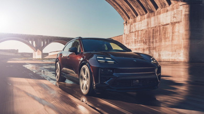 Front three-quarter action shot of a dark bronze Porsche Macan Turbo Electric speeding under a large concrete arched bridge.