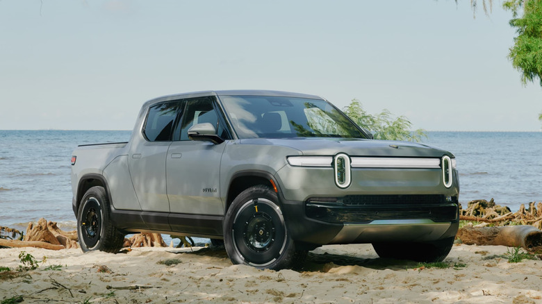 Front three-quarter view of a silver Rivian R1T electric pickup truck parked on a sandy shoreline near the water.