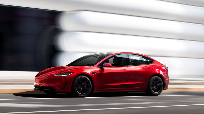 Profile action shot of a red Tesla Model 3 Performance driving at speed past a modern white building.