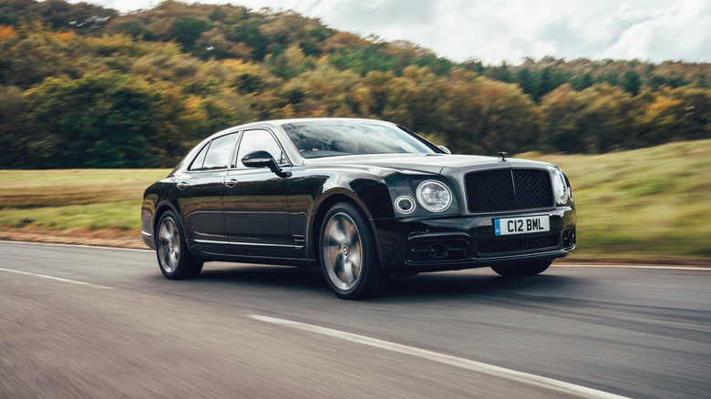 Dark-colored Bentley Mulsanne sedan right front three-quarter view driving on a rural tree-lined road