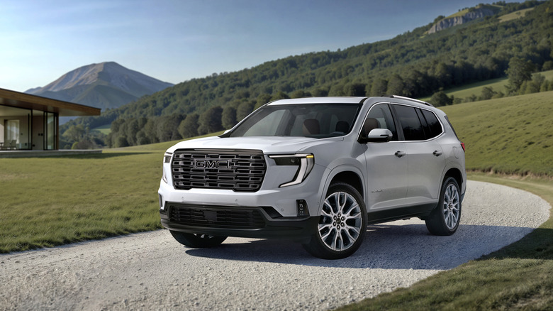 White 2026 GMC Acadia Denali Ultimate SUV left front three-quarter view parked on a gravel road with mountains in background