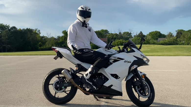 A helmeted rider sits on a white Kawasaki Ninja 400 in a parking lot.