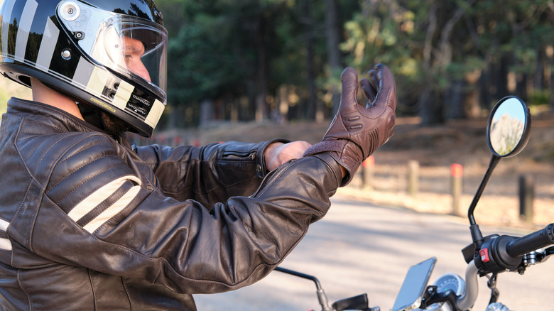 A motorcyclist wearing protective gear sits on the side of road, adjusting a leather glove.