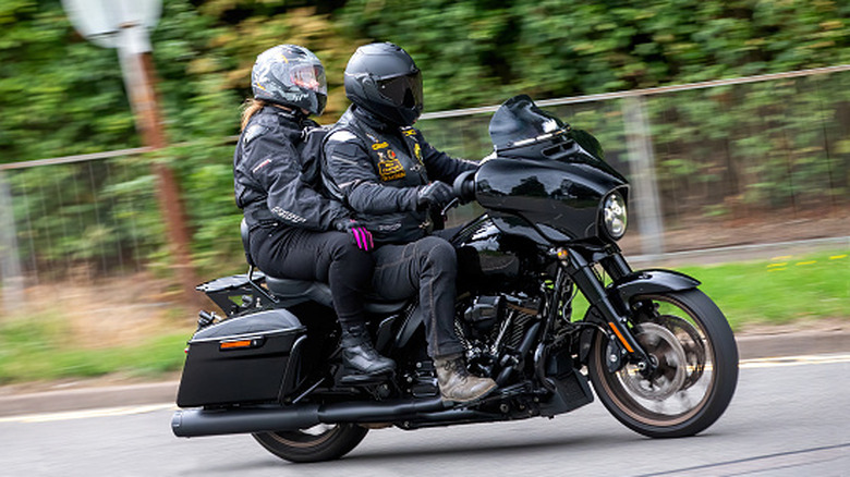 Two motorcycle riders share a bike while outfitted in riding gear.
