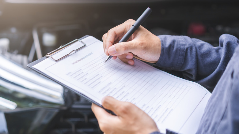 A mechanic writes on a clipboard over an engine bay.