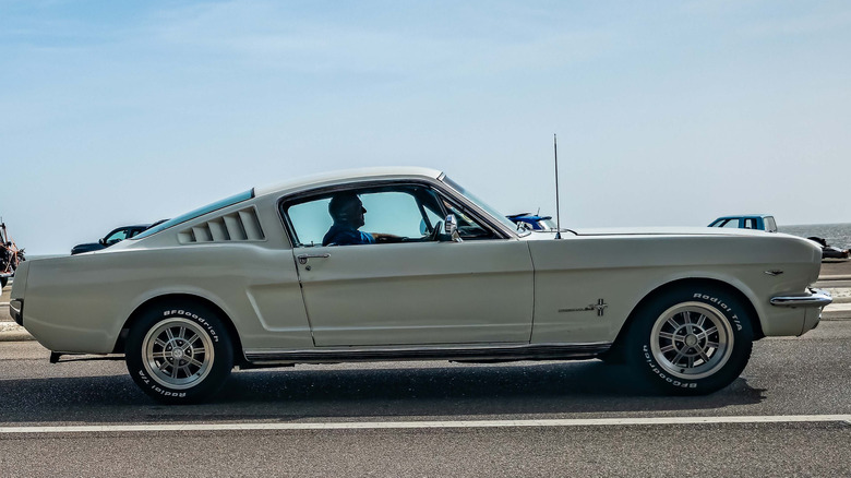 A white 1965 Ford Mustang with a fastback body style driving on the road.