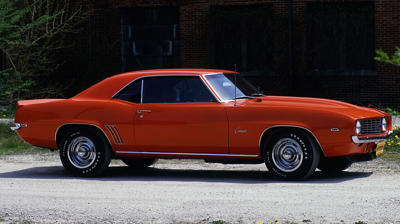 A side view of an orange 1969 Chevy Camaro paked on a gravel road.