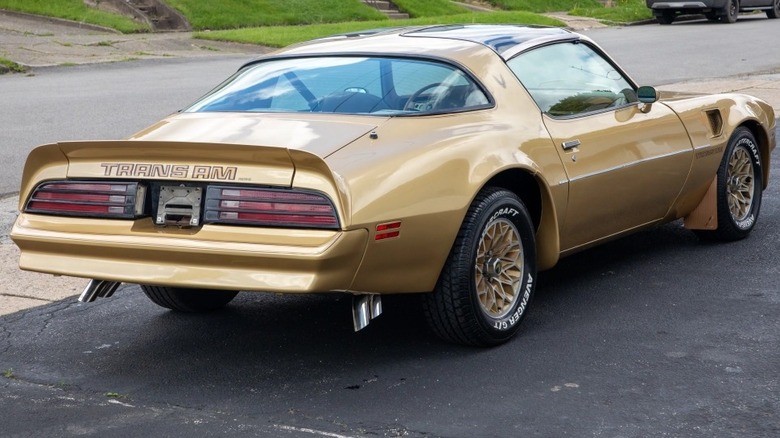 A rear 3/4 view of a 1978 Pontiac Trans Am in gold parked next to a small street.