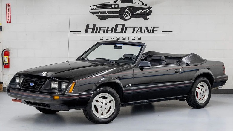 Front three-quarters shot of the black 83 Fox Body parked in a garage with its top down in front of a white wall with the company logo on it