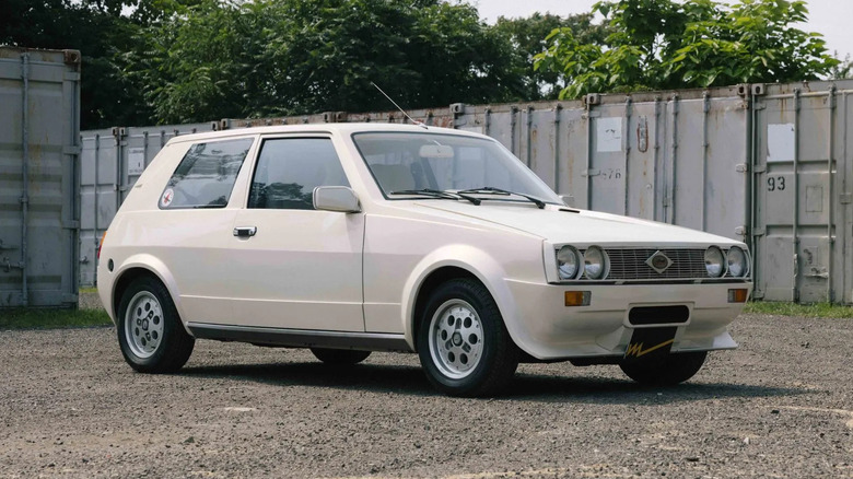 A front three quarters sho of the white Sbarro Super Five parked on dirt in front of gray shipping containers and trees