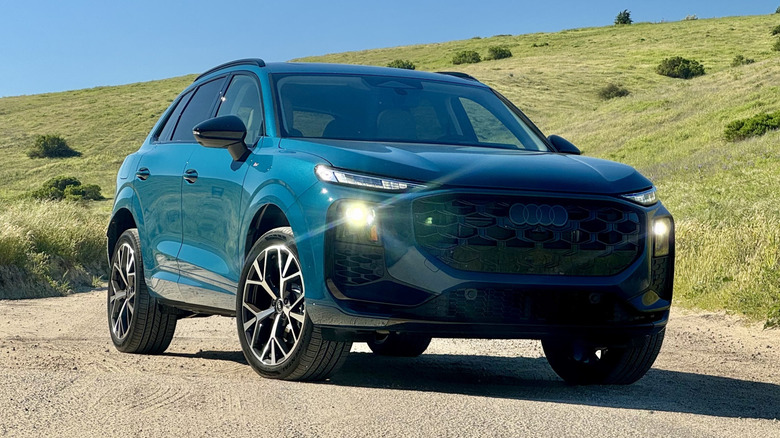 A front three quarters shot of the blue Audi Q3 with its headlights on parked on a dirt road in front of a grass and shrub-covered hill