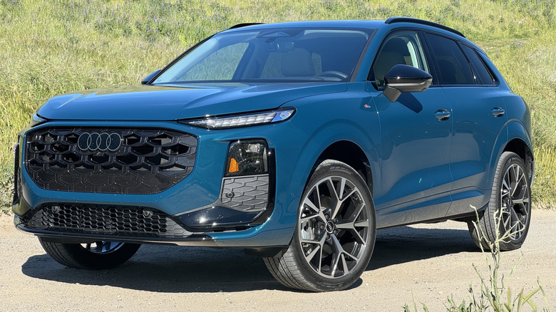 A front three quarters shot of the blue Audi Q3 parked on dirt in front of a grass-covered hill