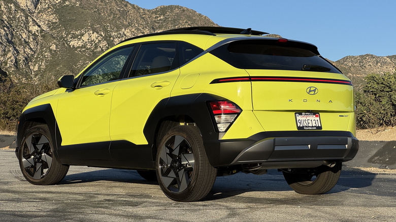 A rear three-quarters shot of the yellow Kona parked on asphalt in front of a shrub-covered mountain