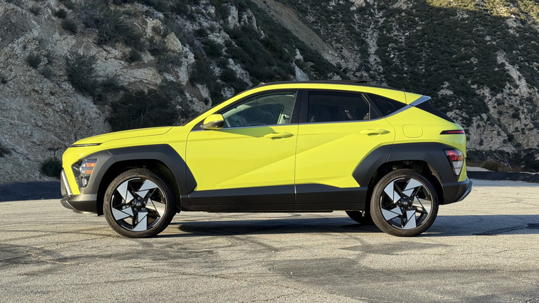 A side view of the bright yellow Kona parked on skid-marked asphalt in front of a shrub-covered mountainside
