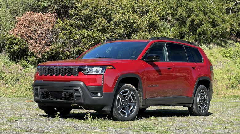 A front three quarters shot of a red Cherokee parked on gravel in front of a shrub-covered mountainside