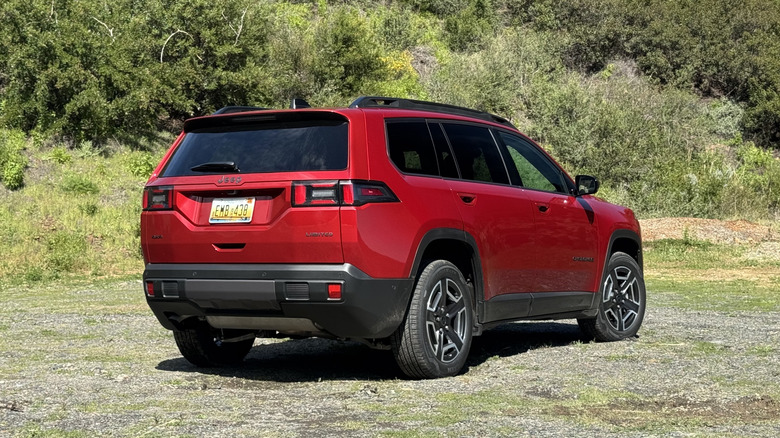A rear three-quarters shot of the red Cherokee parked on gravel in front of a shrub covered mountainside