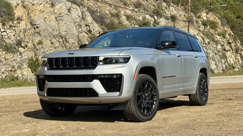Front three quarters shot of a silver Grand Cherokee L parked on dirt in front of a shrubby, rocky mountainside