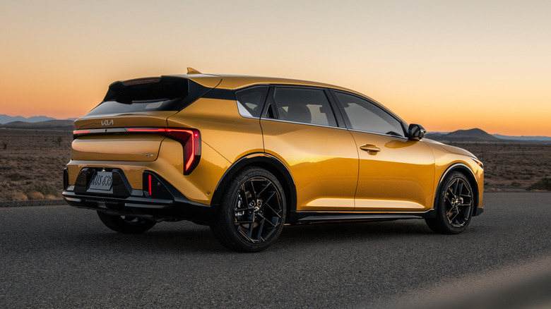 A rear three-quarters shot of a yellow Kia K4 Hatchback parked on asphalt in front of a desert landscape at sunset