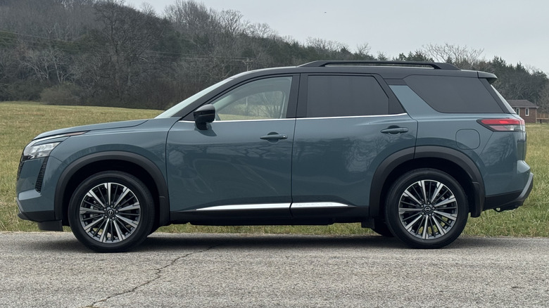 A side view of the teal Pathfinder Platinum parked on asphalt in front of a grassy field and a tree-covered hillside