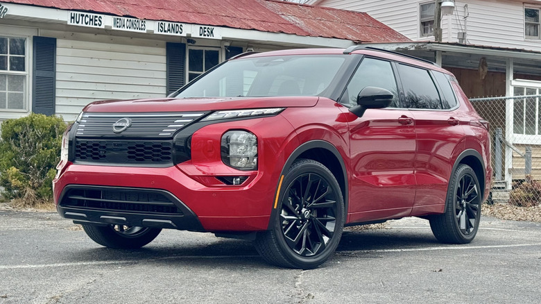 A front three-quarters shot of the red Nissan Rogue PHEV parked in front of a white building advertising different kinds of furniture