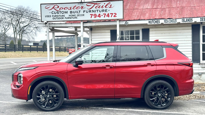A side view of the red Nissan Rogue PHEV parked in front of a white building called Rooster Tails custom built furniture