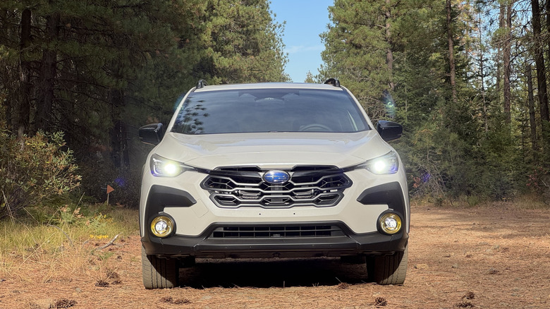 A head-on shot of the white Crosstrek Hybrid with its headlights on parked on pine needles in front of trees