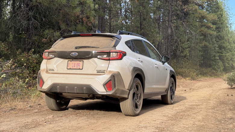 A rear three-quarters shot of the very dusty white Crosstrek Hybrid parked on dirt in front of trees