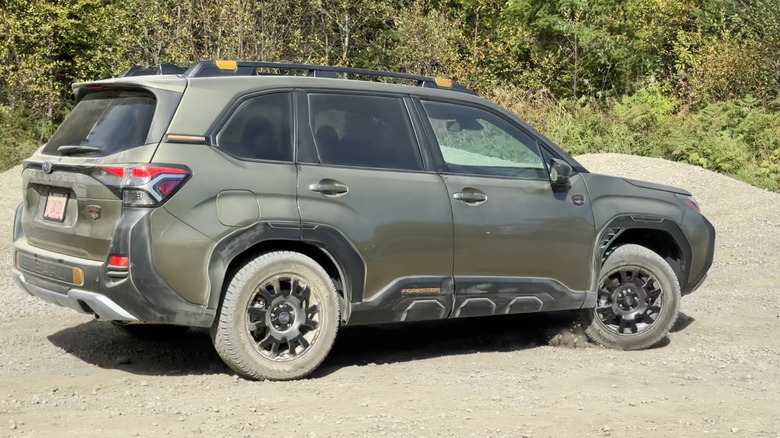 A side view of a green Forester Wilderness driving on gravel in front of trees