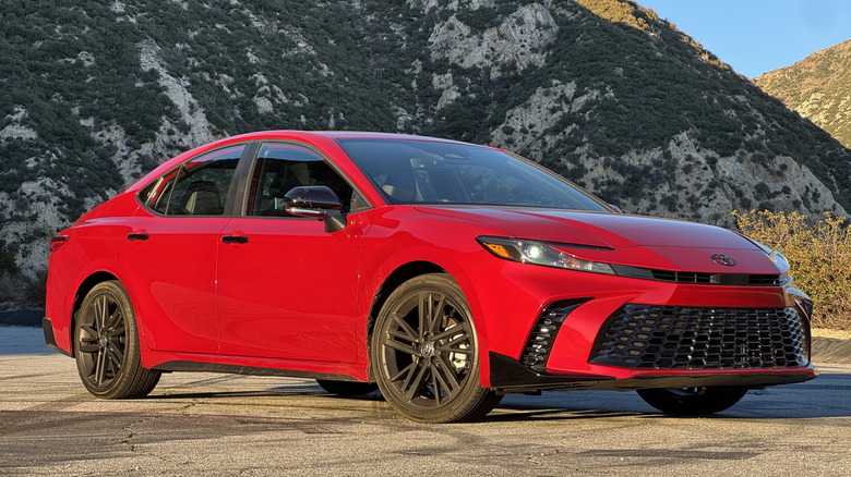 A front three quarters shot of the red Toyota Camry Nightshade Edition parked on asphalt in front of a shrub-covered mountain and a little view of blue sky