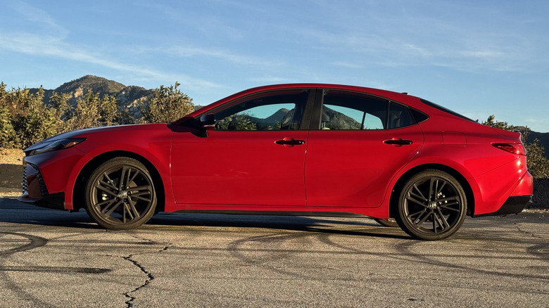 A side view of the red Camry Nightshade parked on asphalt in front of a blue sky, a mountain, and some shrubs
